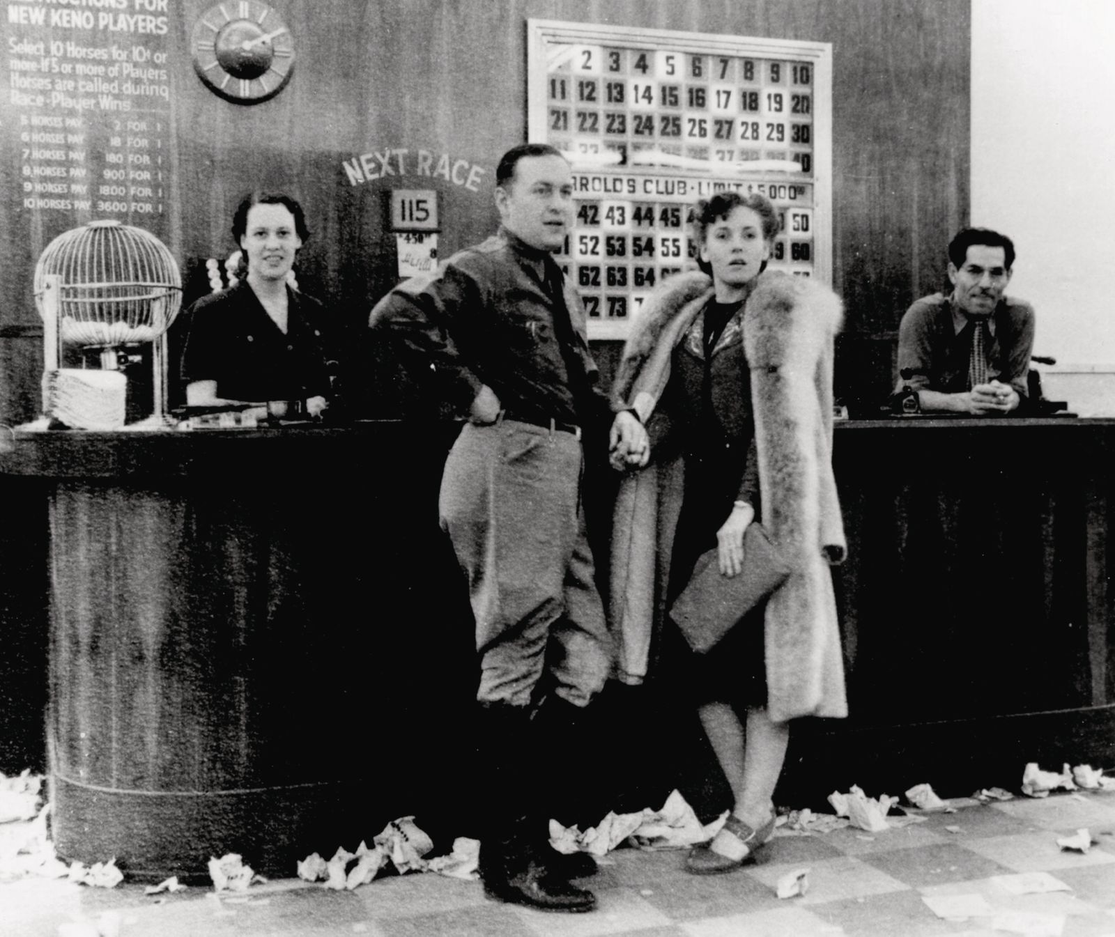A black-and-white photo of a counter in front of which stand a richly-dressed man and woman. Behind the counter are two staff. On the counter is a 'squirrel cage' containing balls. On the wall behind the counter are the rules, a clock, and a board which lights up numbers when they are called.