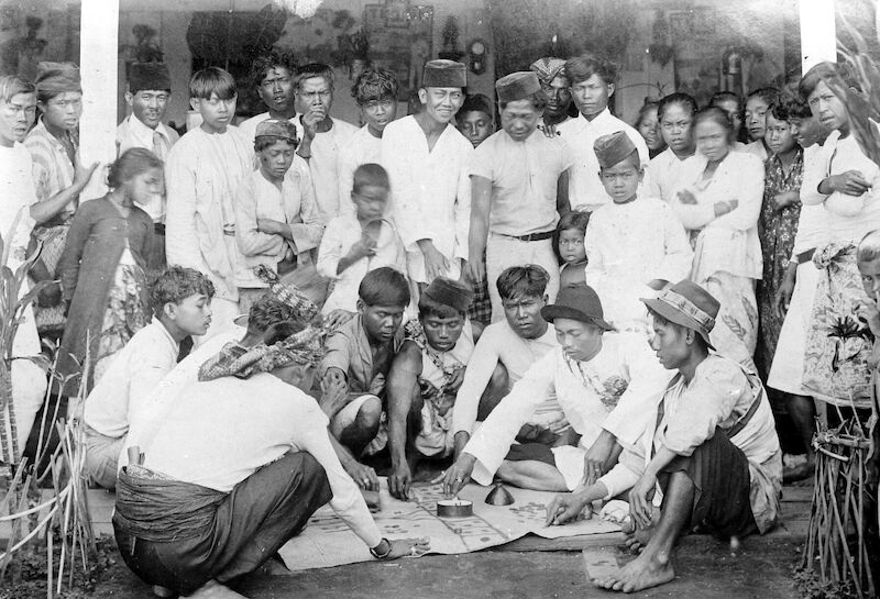 A black-and-white photo of a group of people, mostly men, standing and seated around a mat on the ground. One man is spining a teetotum in a dish, other players are placing coins on the mat inside squares marked with dice symbols.