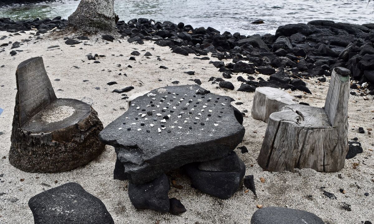 A photograph of a kōnane board set up for play on a beach, with two chairs carved from tree trunks.