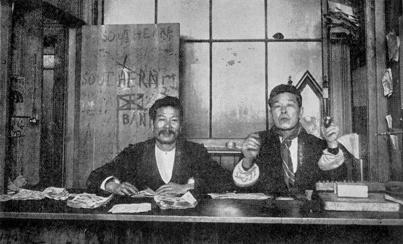 Two Chinese men sitting at a counter in fashionable European suits. The name ‘Southern Bank’ and a rough sketch of a British flag are painted on the door. There are pak-a-poo tickets on the counter. The door reads 'Southern Bank'.