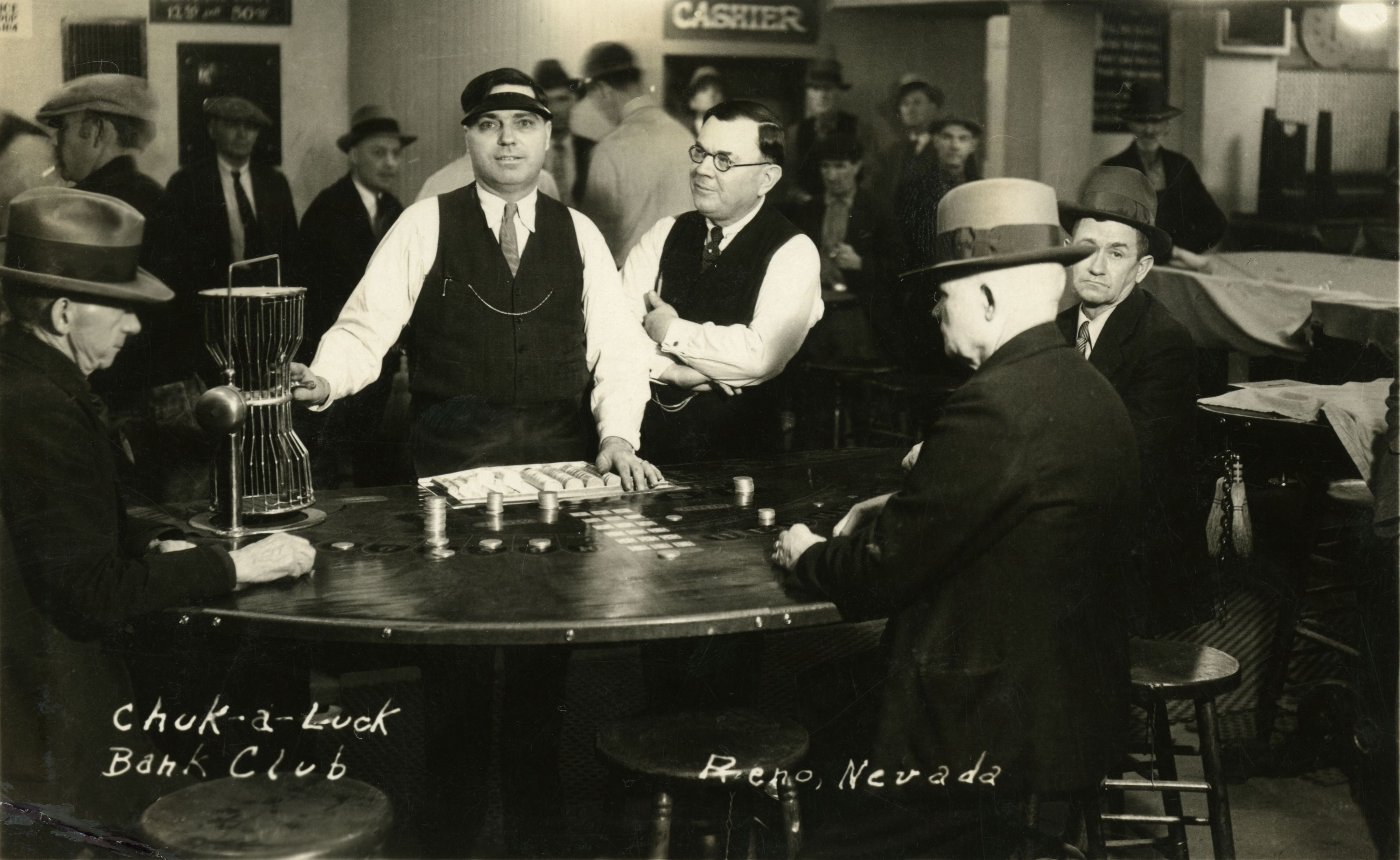 Men sitting around a table with an extended Chuck-A-Luck staking layout and bird-cage sitting on it.