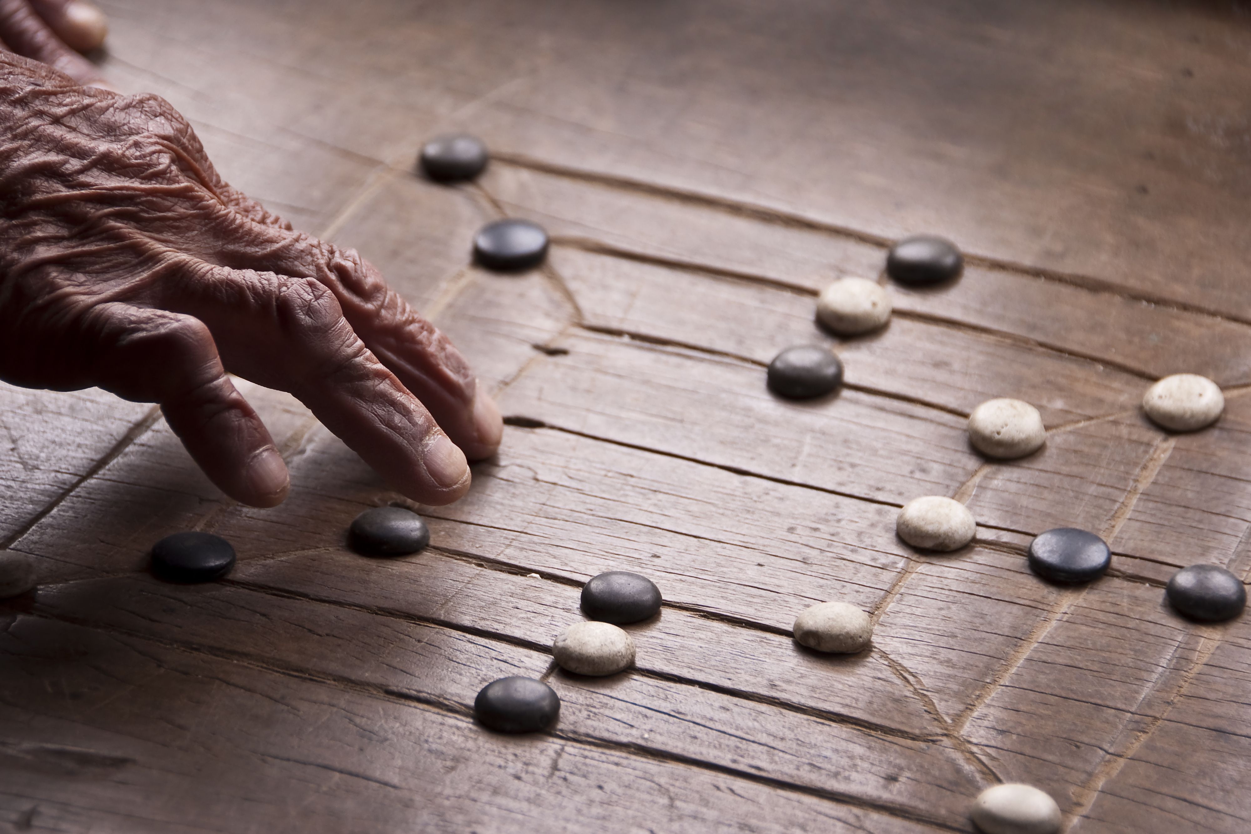 An old manâ€™s hand reaches towards a worn morris board to move a piece.