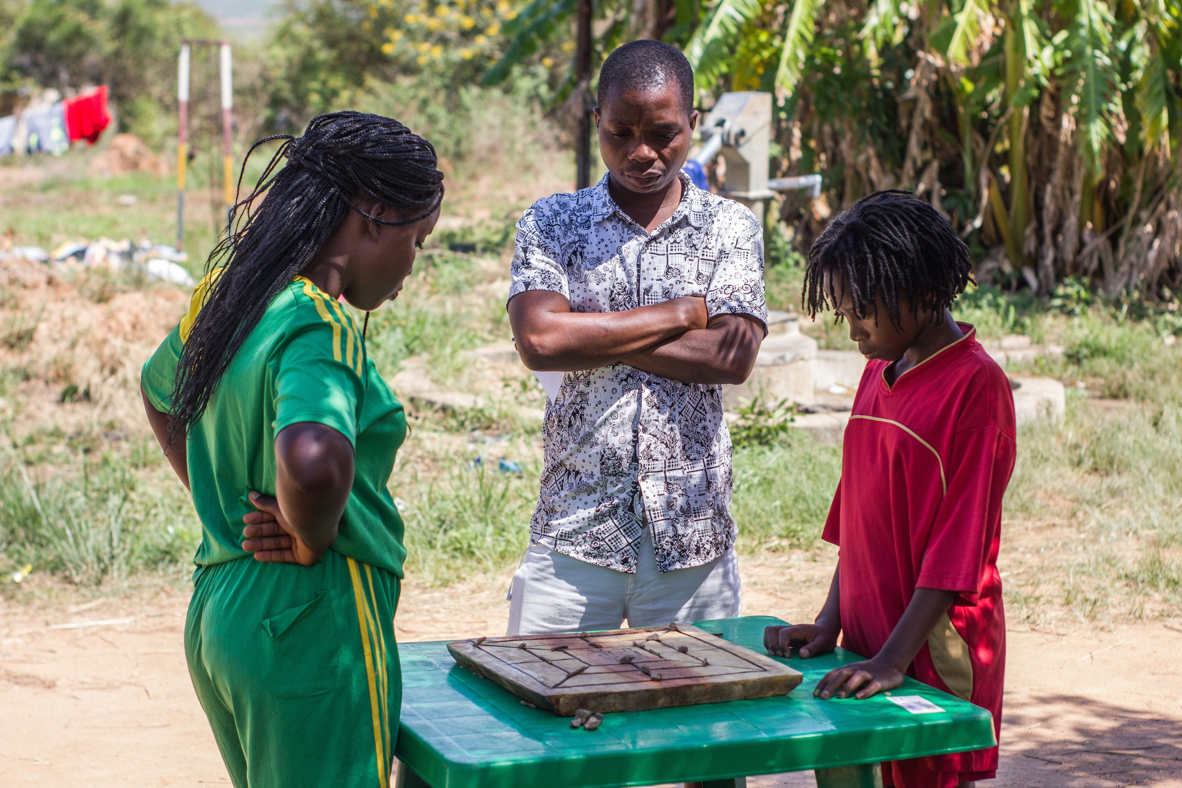 Two school chidren stand next to a table where a game of muravarava is being played, looking at the board intensely, while an umpire watches.