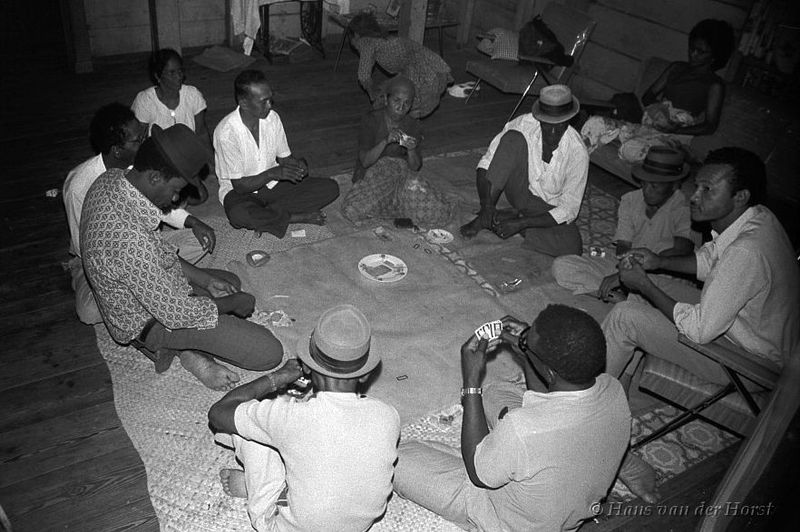 A black-and-white photograph of ten people sitting in a circle on a mat, mostly men, several are wearing pork-pie hats. They are holding cards, and in the centre of the mat is a plate holding ceki cards.