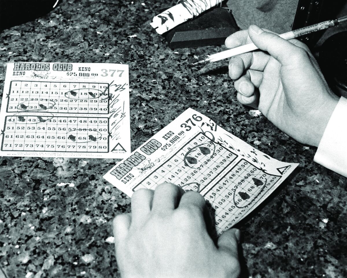 A black-and-white photo of two tickets on a stone counter. Two hands belonging to a man are in the shot: the left is holding the ticket, the right is holding a brush with ink on it.