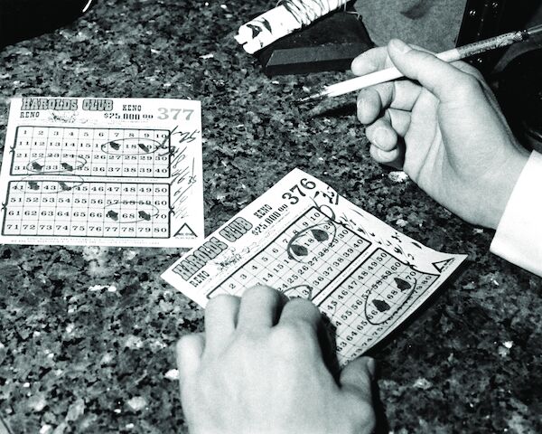 A black-and-white photo of two tickets on a stone counter. Two hands belonging to a man are in the shot: the left is holding the ticket, the right is holding a brush with ink on it.