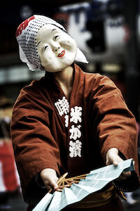 An actor wearing a mask of a white-faced woman with large cheeks, raised eyebrows, and a smile on her lips.