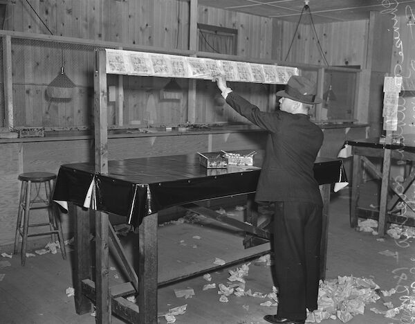 A flash-lit black-and-white photograph of a room. In the centre of the room is a man wearing a suit who is looking at a row of tickets pinned above a table. On the table top are brushes and ink: the table has a heavy plastic tablecloth to protect against ink spillages. On the floor lie many discarded tickets. At the back of the room are a row of teller locations, with low lamps, and protected by chicken-wire mesh.