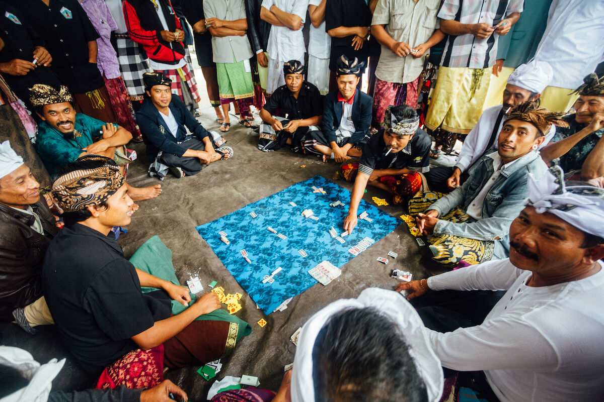 Men in traditional dress seated around a cloth mat upon which are placed ceki cards and bundles of money.