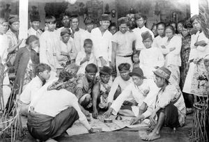 A black-and-white photo of a group of people, mostly men, standing and seated around a mat on the ground. One man is spining a teetotum in a dish, other players are placing coins on the mat inside squares marked with dice symbols.
