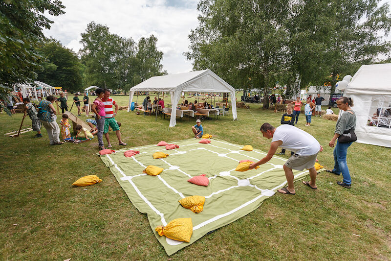 People playing a large nine men’s morris game outdoors on a very large cloth with the board painted on it. They are playing on the grass surrounded by pavilions and onlookers.