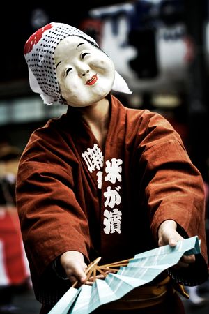An actor wearing a mask of a white-faced woman with large cheeks, raised eyebrows, and a smile on her lips.