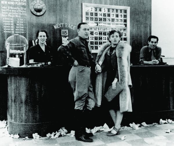 A black-and-white photo of a counter in front of which stand a richly-dressed man and woman. Behind the counter are two staff. On the counter is a 'squirrel cage' containing balls. On the wall behind the counter are the rules, a clock, and a board which lights up numbers when they are called.