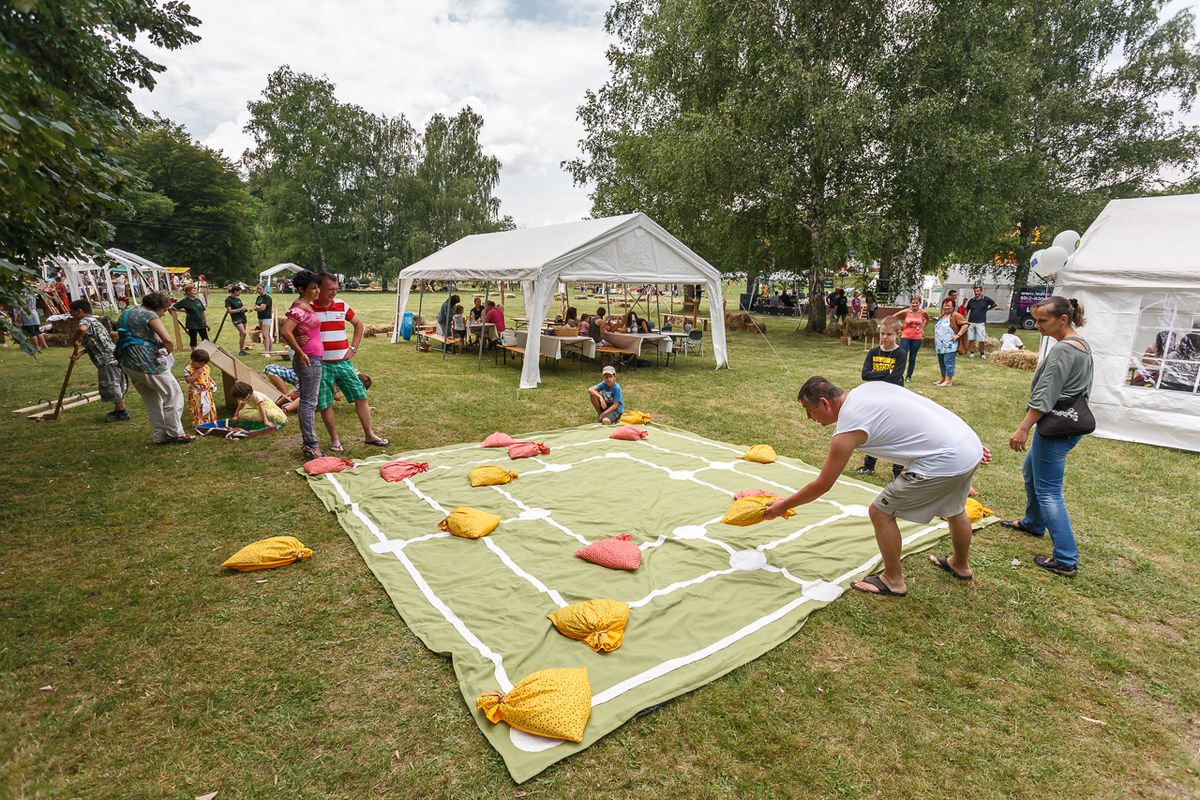 People playing a large nine men’s morris game outdoors on a very large cloth with the board painted on it. They are playing on the grass surrounded by pavilions and onlookers.