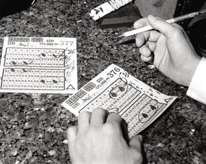 A black-and-white photo of two tickets on a stone counter. Two hands belonging to a man are in the shot: the left is holding the ticket, the right is holding a brush with ink on it.